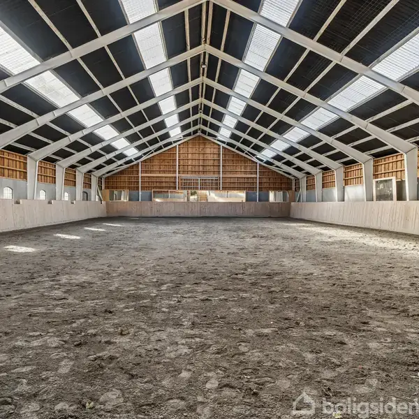 En ridehal med jordgulv står stille, omgivet af høje, hvide trævægge og et buet loft med ovenlys, der giver naturligt lysindfald.