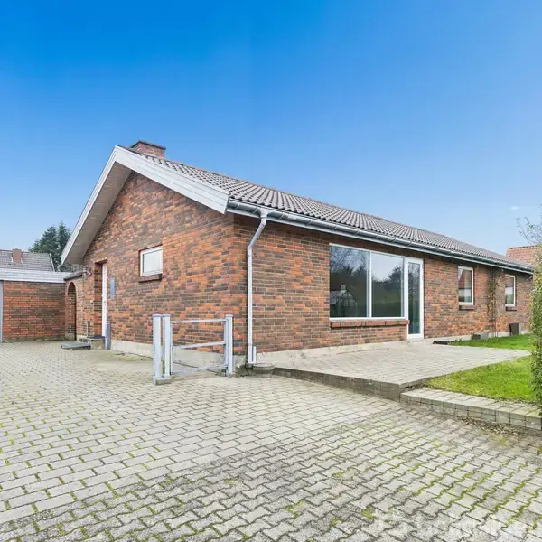 En étplans murstensvilla står stille på en flisebelagt indkørsel ved siden af en garage, omgivet af en velplejet hæk og klar blå himmel.
