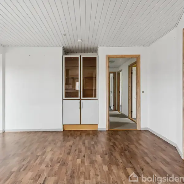 Et tomt rum med trægulv og hvidmalet loft. Skabet i midten står op ad den hvide væg. Trærammede døre fører til andre rum.