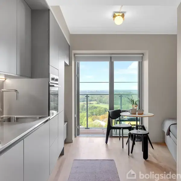 Køkken i moderne stil med grå skabe og stålvaske, åbent til en spiseplads med et bord og stole ved et stort glasvindue med udsigt over grønne landskaber.