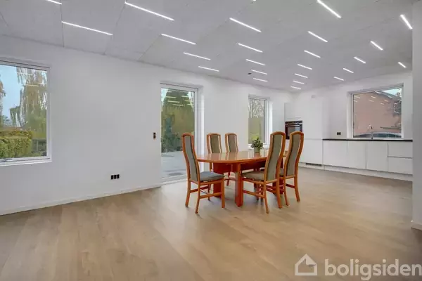 Et spisebord med seks stole står i et lyst rum. Kæmpe vinduer viser natur udenfor. Moderne, minimalistisk indretning med træ- og hvide elementer.