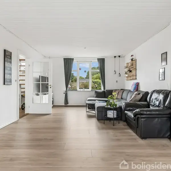 Stue med sort hjørnesofa står mod en hvid væg, dekoreret med billeder og hylder. Store vinduer lader lys ind. Plante i midten på et lille bord. Trægulv.