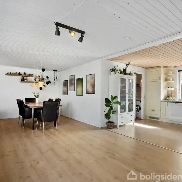 Spisebord med sorte stole står i et lyst rum med trægulv. Bordet er dækket af planter. Til højre ses et køkken med hvide skabe og et vindue.