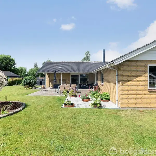 Étplanshus med gult murværk står i en velplejet have. Terrassen foran huset er indrettet med havemøbler. Træer og buske omgiver området, himlen er overvejende klar.