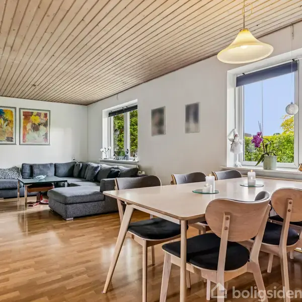 En stue med en grå hjørnesofa står på et trægulv. Til højre er der et spisebord med seks stole. Store vinduer slipper naturligt lys ind, og der hænger kunst på væggene.