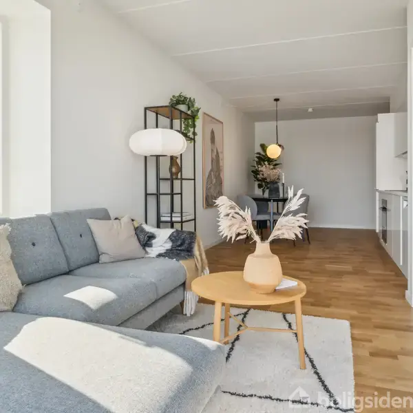 Grå hjørnesofa findes i en lys stue med trægulv, dekoreret med puder og tæppe. Træbord med vase i forgrunden; i baggrunden ses spisebord under loftslampe.