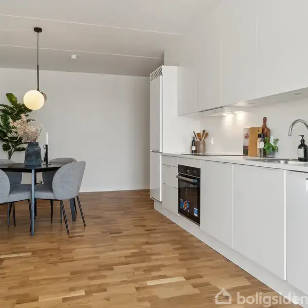 Moderne køkken-alrum med hvide skabe, indbygget ovn og sort bordplade. Rundt spisebord med grå stole står i hjørnet ved en stor plante og hængelampe. Trægulv.