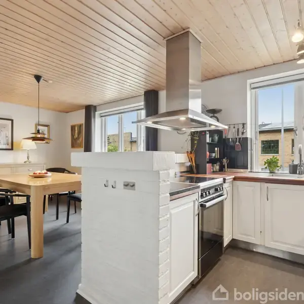 Køkken med en moderne ovn og emhætte, træloft og hvide skabe. Til venstre ses et spisebord med sorte stole i en lys spisestue. Ingen tekst til stede.