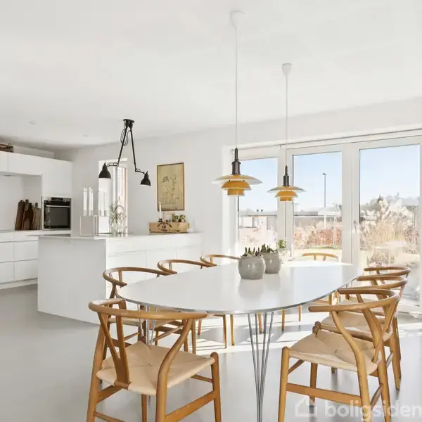 Ovalt spisebord med træstole står under to hængende lamper i et lyst, moderne køkken med hvide skabe og en ø afrustede apparater. Vinduer viser en udenforliggende have.