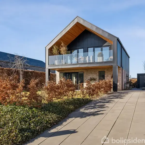 En moderne to-etagers villa med træbeklædt altan står for enden af en flisebelagt indkørsel. Omgivet af vinterklar hæk og græsplæne, postkassen viser nummer "18".