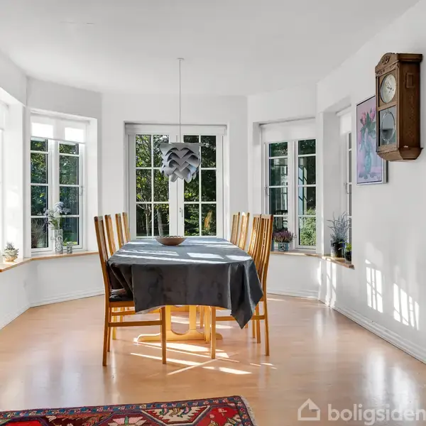 Et spisebord med seks træstole står centralt i et lyst rum. Bordet er dækket med en grå dug. Store vinduer omkranser rummet, der har en vægmonteret pendel og et ur.