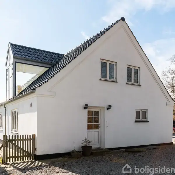 Hvidt hus står stille på en grusvej omgivet af træer og græsmarker. Huset har mørkt tag og flere vinduer, inklusive en glasudbygning på første sal.