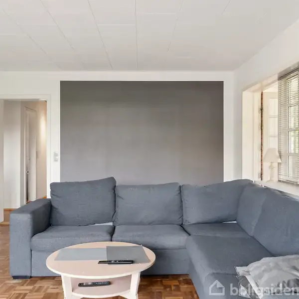 Grå sofa står i en stue foran en sort væg. Rundt sofabord med fjernbetjening. Store vinduer til højre med persienner, træparketgulv. Døråbning til venstre side.