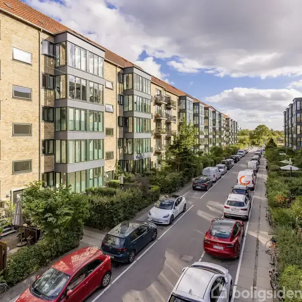 Flere etagebygninger står langs en gade fyldt med parkerede biler. Bygningerne har moderne, glasindfattede altaner. Gaden er omgivet af grønne planter og træer under en blå og delvist overskyet himmel.