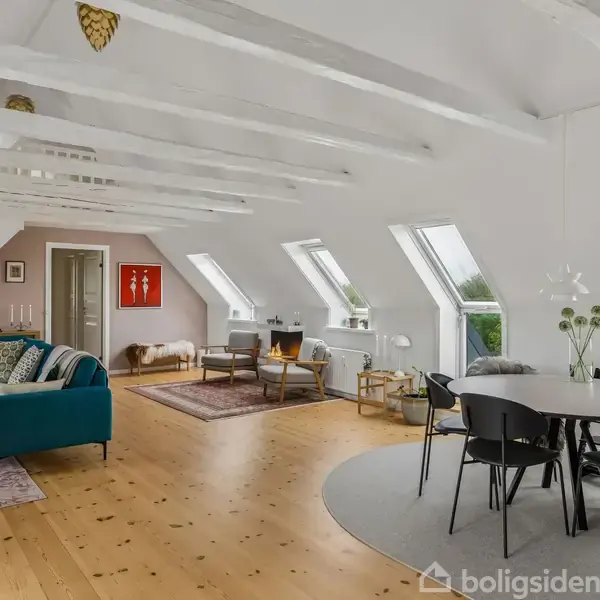 Stue med blå sofa og spisebord, placeret under loftsbjælker i et lyst rum med skråvinduer. Trappe i baggrunden fører til overetagen. Planter og kunstværker dekorerer væggene.
