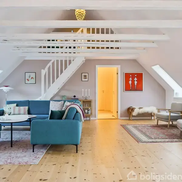 Stue med blå sofa og sofabord under loftsbjælker, trappe fører til første sal. Rummet har skråvinduer og trægulv, dekoreret med kunst, hvide møbler og tæpper.