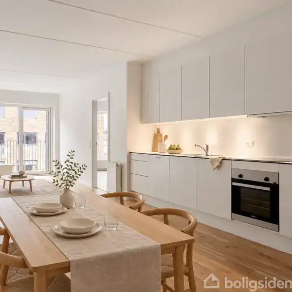 Spisebord med dækket service i et lyst køkkenalrum. Køkkenet har hvide skabe, træbord, og stor vinduesparti ved siden af en grå sofa. Væggen til venstre har et åbent rum.