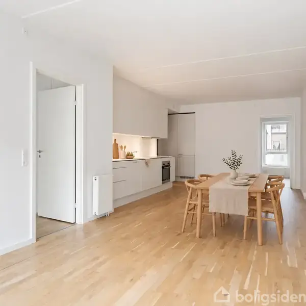 Køkken-alrum er enkelt med træspisebord, fire stole og en beige dug. Hvide køkkenskabe og en komfurzone findes bagerst, mens rummets vægge er hvidmalede.