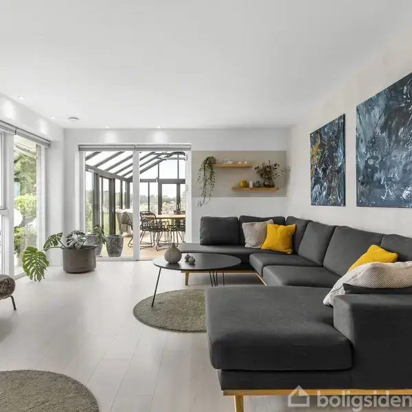 En stue med en stor grå hjørnesofa, dekoreret med gule puder, står mod en hvid væg med kunstværker. Rummet har store vinduer mod en udestue med planter og spisebord.