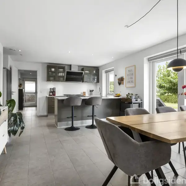 Spisestue og køkken med træbord og grå stole. Køkkenet har en bar med to stole. Lyse vægge, store vinduer med udsigt til grøn natur og moderne indretning. Ingen tekst.
