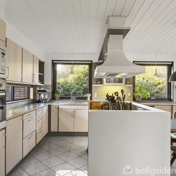 Et moderne køkken med lyse trækøkkener og stålapparater. Det har en stor emhætte over en ø. Der er store vinduer med udsigt til grønne planter udenfor. En spiseplads med en skål appelsiner fuldender rummet.