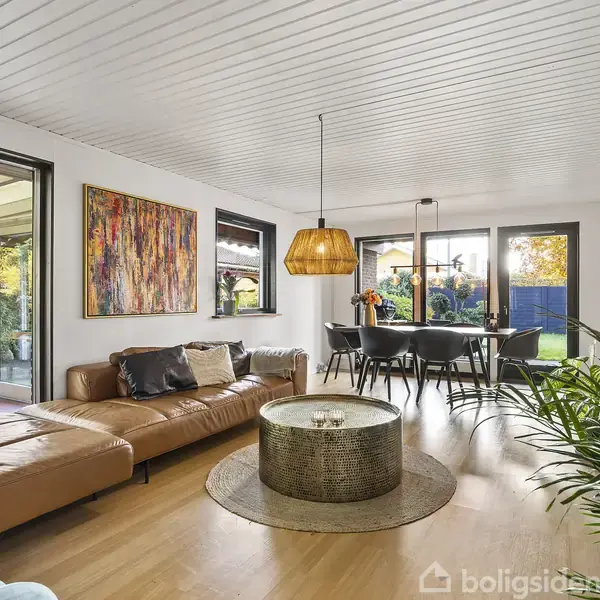 Lædersofa står i en stue ved siden af et stort, rundt sofabord. En lampe hænger over spisebordet i nærheden, med udsigt til have gennem store vinduer. Maleri hænger på væggen.