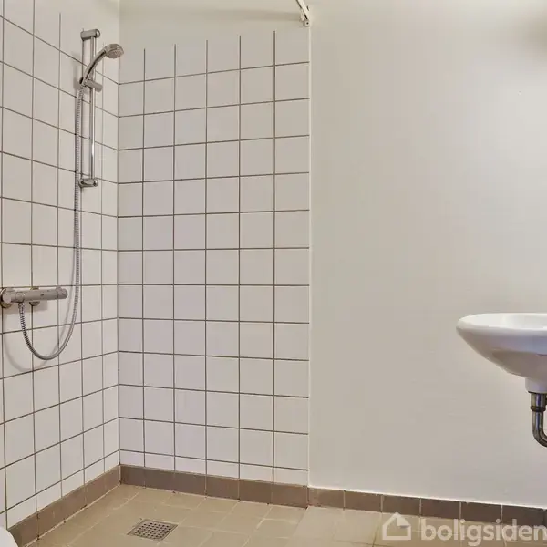 Bruser og håndvask er fastgjort på væggen i et badeværelse med hvide fliser og brune gulvklinker. Miljøet er minimalistisk og funktionelt.