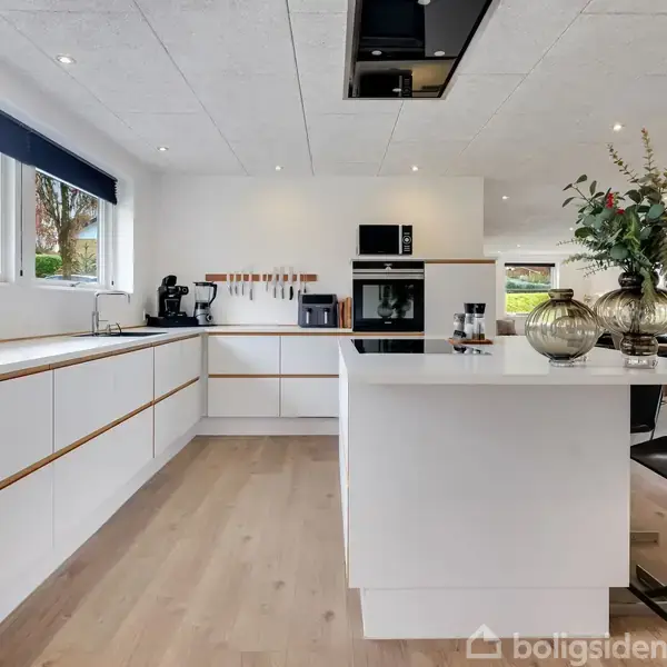 Et moderne køkken med hvide skabe og en stor ø. På bordet står dekorative glasvaser med planter. Store vinduer giver naturligt lys. Indgangen til spisestue er synlig.