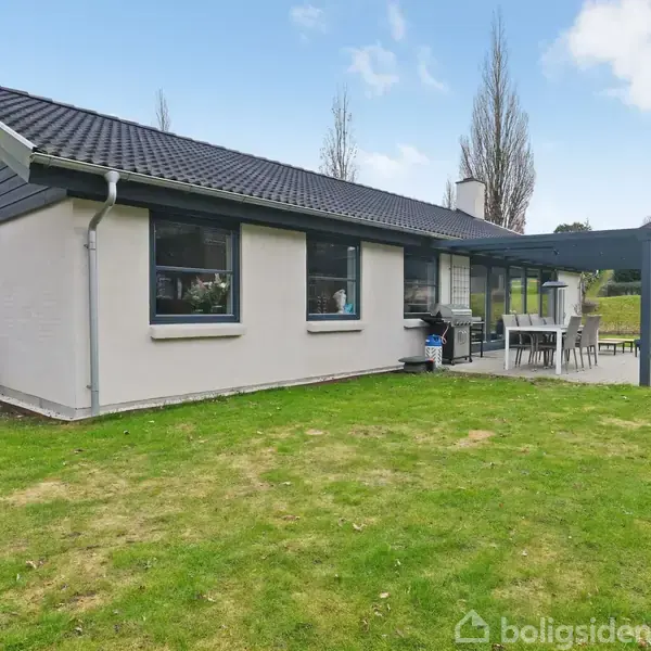 En enkelthus med hvide vægge og gråt tag står stille i en grøn have. Terrassen ved siden af er møbleret med spisebord og grill under en pergola.