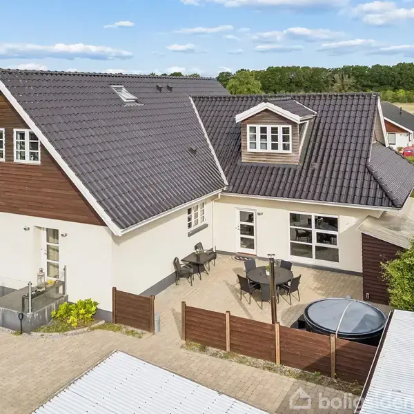 Huset er et moderne to-etagers hjem med mørkt tegltag. En terrasse med bord og stole ses foran, omgivet af lavt hegn. Omgivelserne inkluderer andre huse og grønne områder.