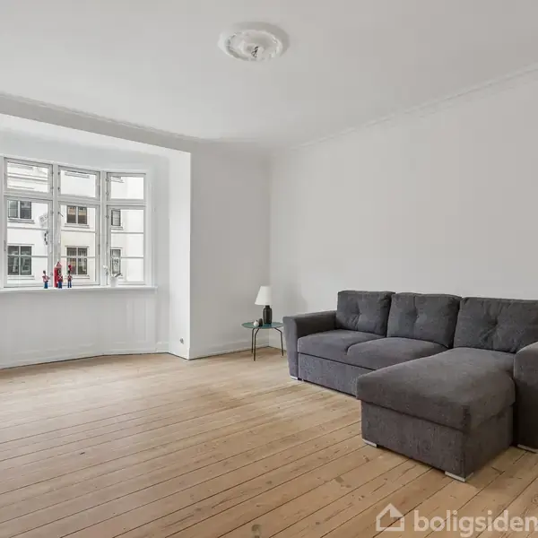 Grå sofa står i en lys stue med trægulv. Vindue med udsigt til bygninger udenfor. Et bord med lampe står ved siden af sofaen. Ingen tekst i billedet.