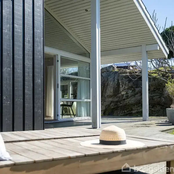 En træterrasse med en solhat placeret på brædderne foran et moderne, sort sommerhus. Terrassen er delvist overdækket, og en grøn have samt klippeformationer ses i baggrunden.