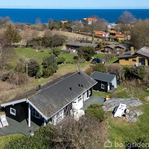 Et fritidshus ligger i en grøn have med en træterrasse og hvide liggestole. Flere huse og træer omkranser området, mens havet er synligt i baggrunden.