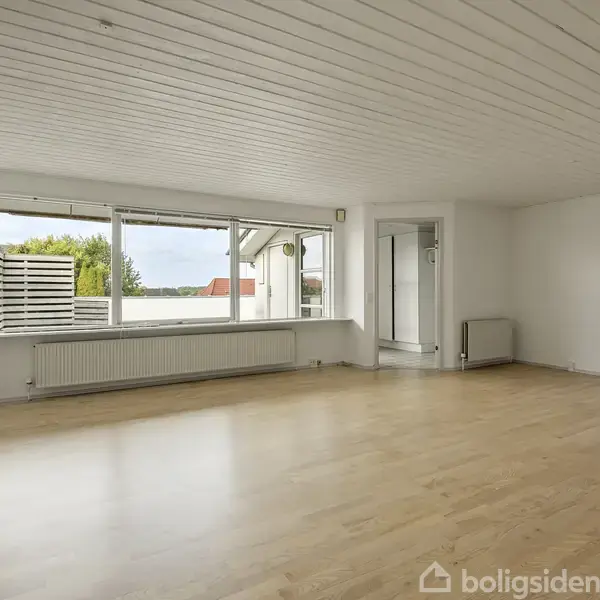 Et rummeligt, lyst værelse med trægulv og hvidmalet loft. Store vinduer giver udsigt til en terrasse og natur udenfor. Der er en radiator under vinduerne og en dør til højre.