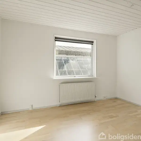 Et tomt rum med trægulv, hvidmalede vægge og loft. En radiator er placeret under et vindue med udsigt til en glasoverdækket struktur udenfor. Ingen tekst til stede.