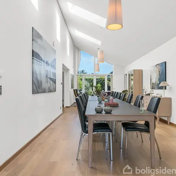 Langt spisebord med sorte stole er centreret i et lyst rum. Store vinduer og skråt loft med hængende lamper tilføjer en elegant atmosfære, omkranset af hvide vægge og træmøbler.