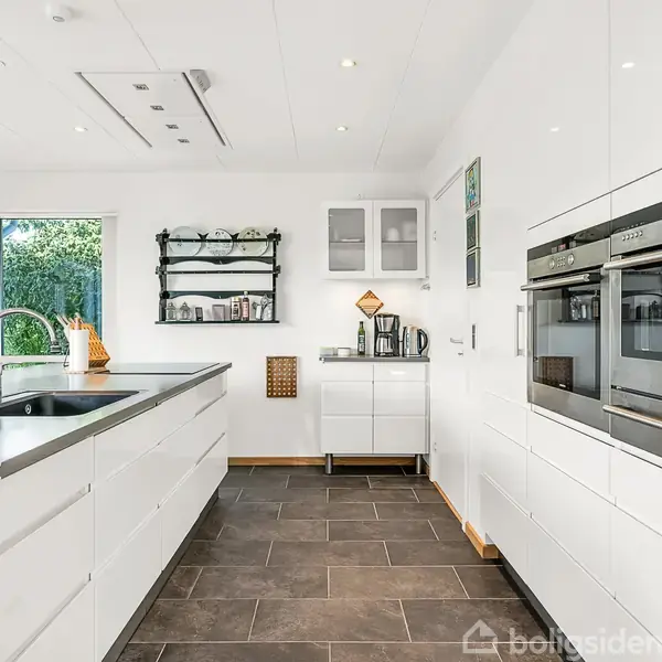 En moderne, hvidt køkken med en stor ø i midten og vask. Væggen til højre har indbyggede ovne. Rummet har mørke flisegulve og åbner ud mod en have.