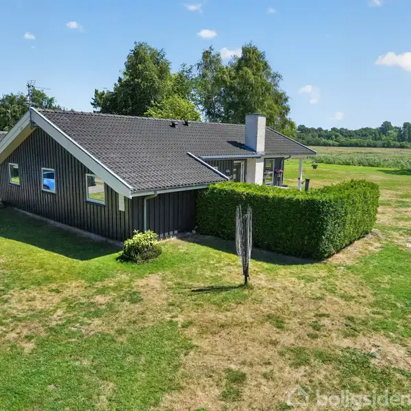 Et sort, moderne hus med skråt tag står på en stor, grøn græsplæne. Omkranset af træer og buske i en landlig, åben natur.
