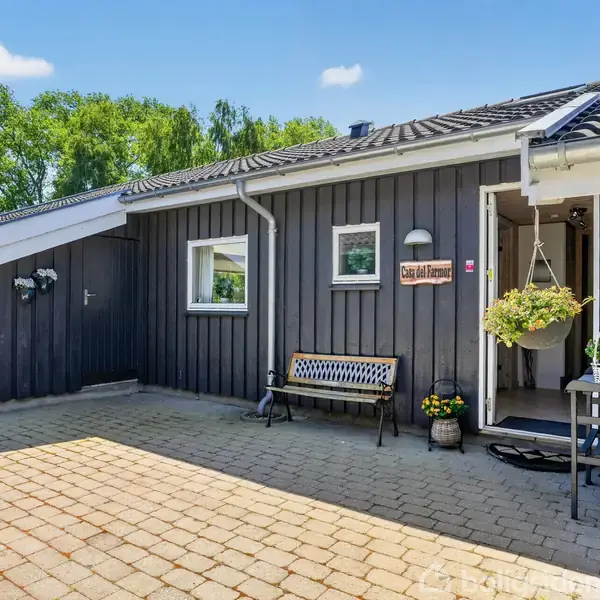 Hus med sort facade har en gårdsplads med fliser. En bænk står ved siden af et skilt med teksten "Guds Aftengrøde". Området er omgivet af grønne træer under blå himmel.