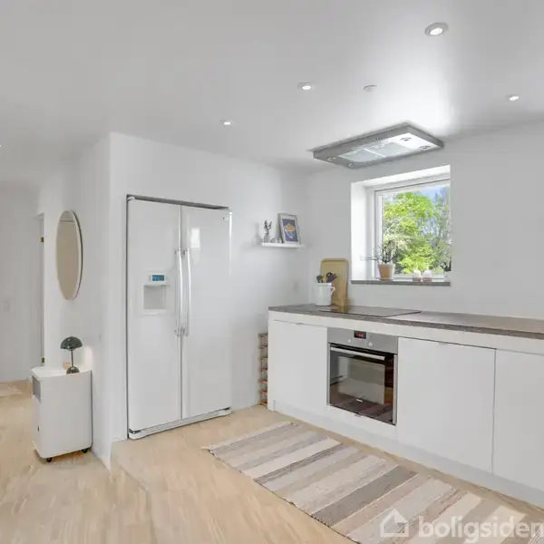 Køkken med hvide skabe og indbygget ovn, stående ved siden af stort hvidt køleskab; enkel indretning med trægulv og to vinduer. Hylde med dekorative genstande over køkkenbordet.