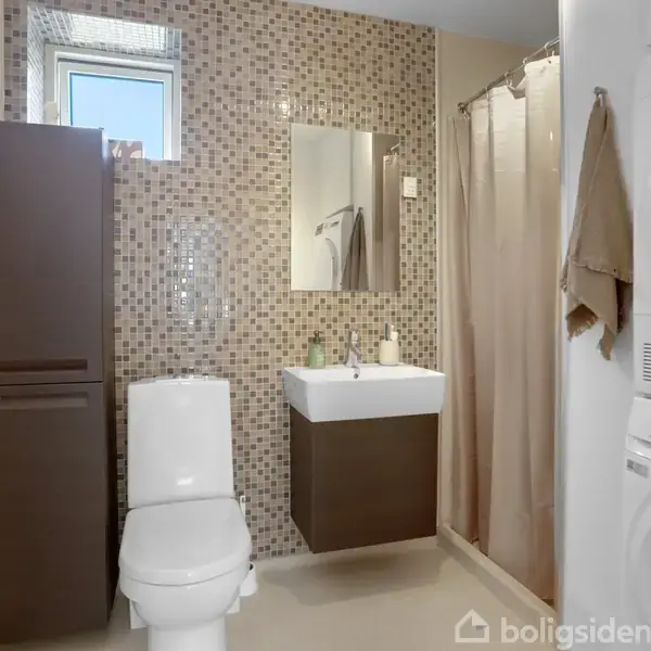 Toilet i et badeværelse med mosaikvæg, bruseforhæng, håndvask med spejl, og vaskesøjle til højre. Skab ved siden af toilettet, vindue over. Ingen tekst til stede.