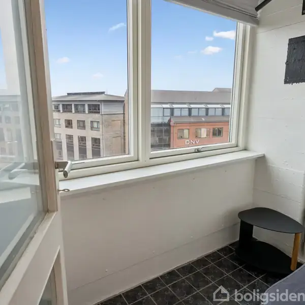 Inderglaset altan med sorte fliser og en lille sort bord. Udsigt gennem store vinduer til bybygninger, herunder en med "DNV" logoet.