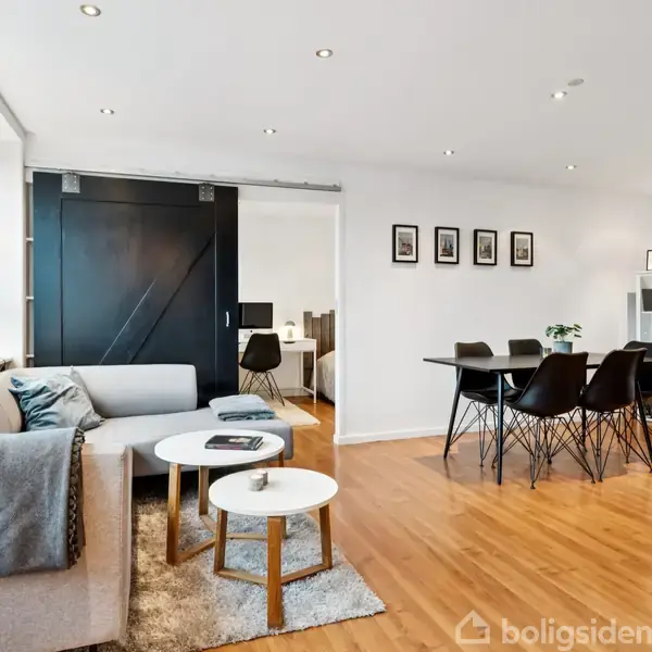 Stue med en lys sofa og to runde borde, placeret på et tæppe. Spisebord med sorte stole står ved siden af en skænk. Hvide vægge har indrammede billeder.