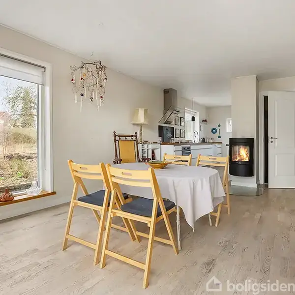 Et spisebord med gule stole står i en lys stue. Et tæppe ligger på gulvet. Vindue viser have. En brændeovn brænder ved siden af køkkenet i baggrunden.