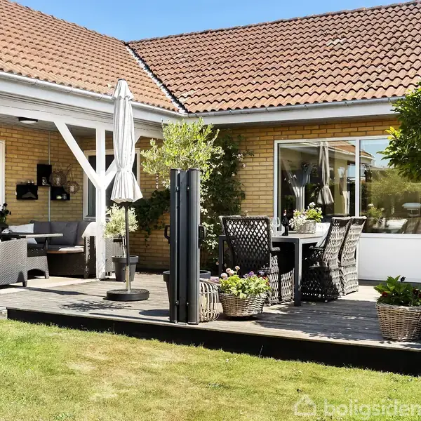 Rækkehus med rød tegltag og terrasse m. havemøbler, omkranset af grønne buske og blomsterkrukker. Terrassebord med stole og parasol ses ved siden af havedør, under blå himmel.