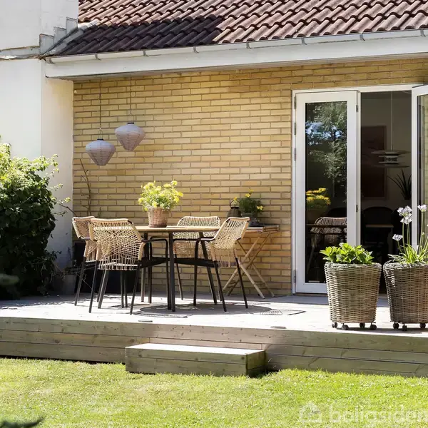 En terrasse med bord og fire stole er placeret udenfor et gult murstenshus med hvide vinduer. Omgivet af grønne planter i krukker og en velplejet græsplæne.