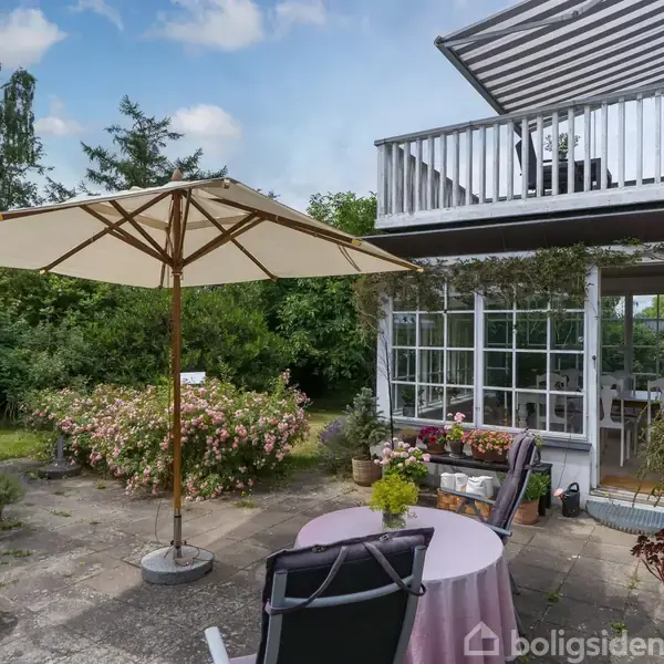 En parasol står på en terrasse med bord og stole, omgivet af planter og blomster. Et hvidt hus med glasveranda ses i baggrunden, og himlen er blå med skyer.