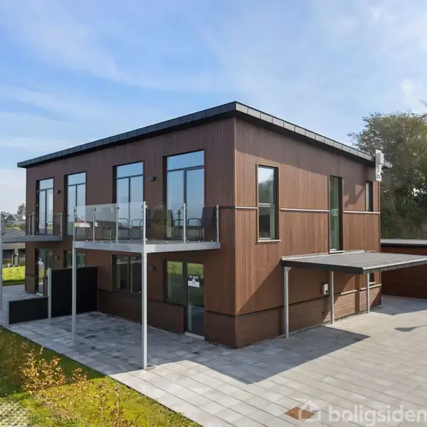 To-etagers moderne hus med store glasvinduer omgivet af flisebelagt terrasse og grønne områder. Huset har en altan og ligger i et beboelseskvarter med andre bygninger i baggrunden.