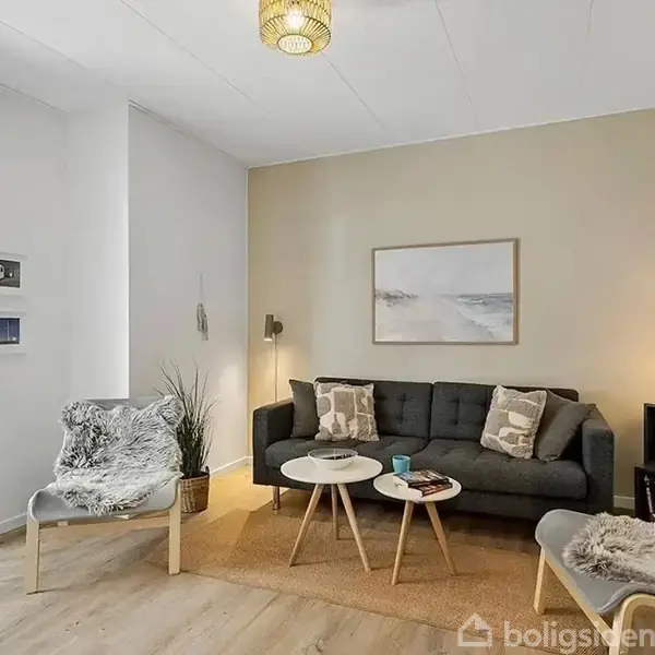 En moderne stue med en grå sofa og to stole med pelsbetræk placeret omkring runde sofaborde. En lampe hænger fra loftet, og et maleri pryder væggen.