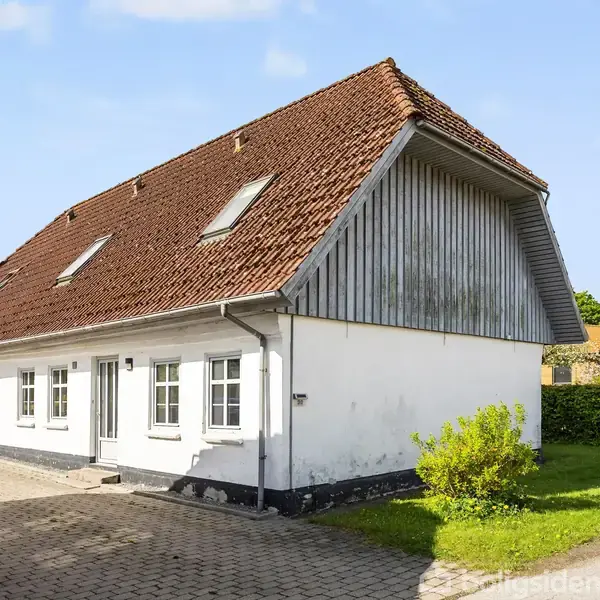 Hus med hvid facade og rødt tegltag står frit, omgivet af have og flisebelagt indkørsel. Der er grønne buske og en blå himmel i baggrunden.